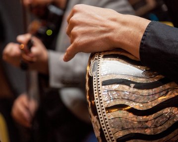 A hand is playing on a drum with a decorative surface, while in the background, a musician with a violin can be seen.