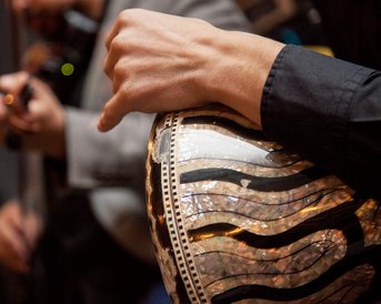 A hand is playing on a drum with a decorative surface, while in the background, a musician with a violin can be seen.