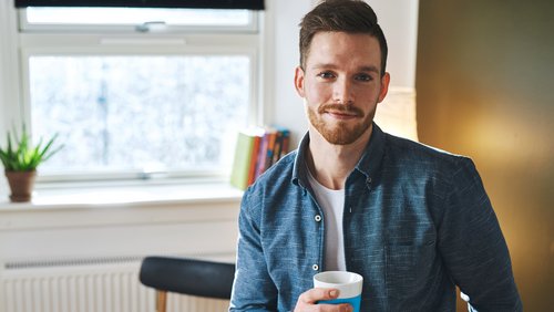 Ein Mann mit Bart hält eine Tasse in der Hand und lächelt. Er trägt ein blaues Hemd und steht in einem hellen Raum mit Pflanzen im Hintergrund.