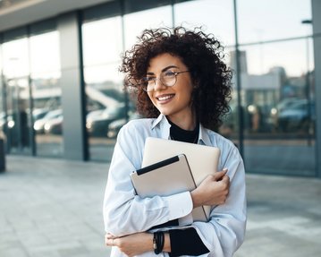 A smiling woman with curly hair is holding a laptop and a notebook, standing in front of a modern building with large windows.