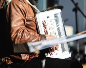 A musician is playing an accordion while sitting on a chair. He is wearing a brown leather jacket and has a scarf around his neck.