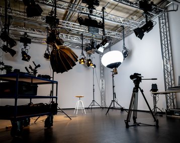 Ein Studio mit verschiedenen Beleuchtungseinrichtungen, Kameras und einem Stuhl in der Mitte. Im Hintergrund sind Stative und technische Ausrüstung sichtbar.