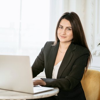 Eine Frau in einem schwarzen Anzug sitzt an einem Tisch mit einem Laptop. Sie schaut direkt in die Kamera, während Tageslicht durch ein Fenster strömt. Pflanzen sind im Hintergrund sichtbar.