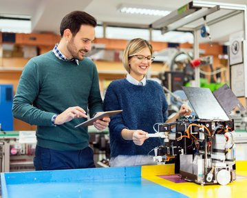 Two people in a lab are working on a technical device, with one holding a tablet and the other inspecting the device.