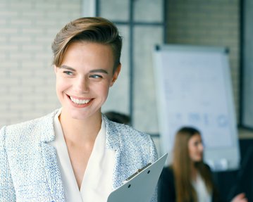 A smiling woman in a blue blazer is holding a clipboard while standing in a modern office. In the background, other people can be seen.