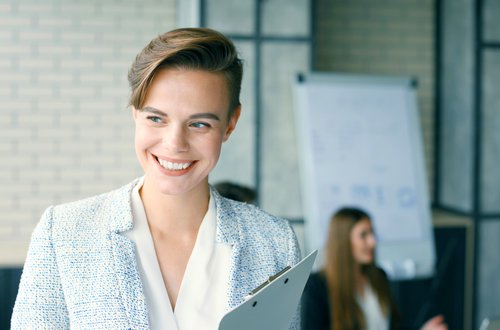 Eine lächelnde Frau in einem blauen Blazer hält eine Clipboard in der Hand, während sie in einem modernen Büro steht. Im Hintergrund sind weitere Personen zu sehen.