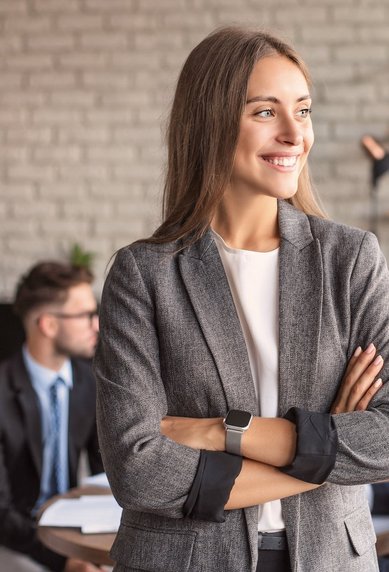 Eine lächelnde Frau in einem grauen Blazer steht mit verschränkten Armen in einem modernen Büro, während im Hintergrund mehrere Personen an einem Tisch diskutieren. Pflanzen und eine helle Wand schaffen eine angenehme Atmosphäre.