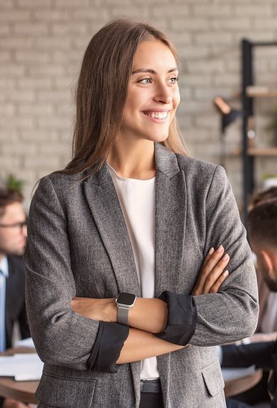 Eine lächelnde Frau in einem grauen Blazer steht mit verschränkten Armen in einem modernen Büro, während im Hintergrund mehrere Personen an einem Tisch diskutieren. Pflanzen und eine helle Wand schaffen eine angenehme Atmosphäre.