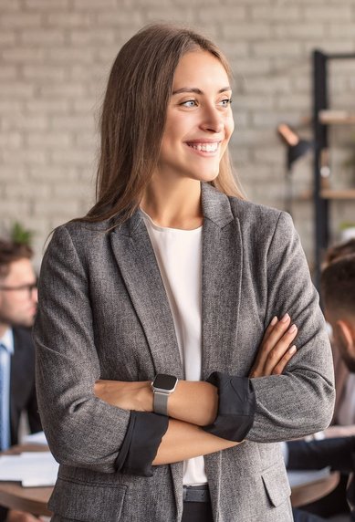 Eine lächelnde Frau in einem grauen Blazer steht mit verschränkten Armen in einem modernen Büro, während im Hintergrund mehrere Personen an einem Tisch diskutieren. Pflanzen und eine helle Wand schaffen eine angenehme Atmosphäre.