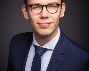 Ein junger Mann in einem eleganten Anzug mit Krawatte und Brille, lächelt freundlich vor einem dunklen Hintergrund.