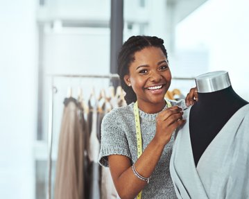 Eine lächelnde Frau mit Zopf steht neben einer Schneiderpuppe und passt ein graues Kleid an. Im Hintergrund hängen Kleider auf Kleiderständern.