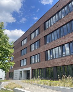 Modernes Gebäude mit roten Ziegeln und großen Fenstern, umgeben von Bäumen und einer gepflasterten Fläche.