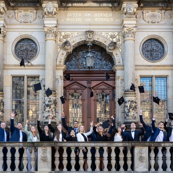 Gruppe von Absolventen in formeller Kleidung, die ihre Hüte vor einem historischen Gebäude in die Luft werfen.