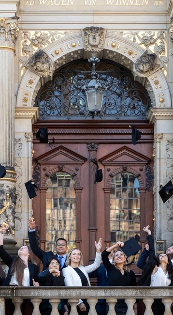 Gruppe von Absolventen in formeller Kleidung, die ihre Hüte vor einem historischen Gebäude in die Luft werfen.