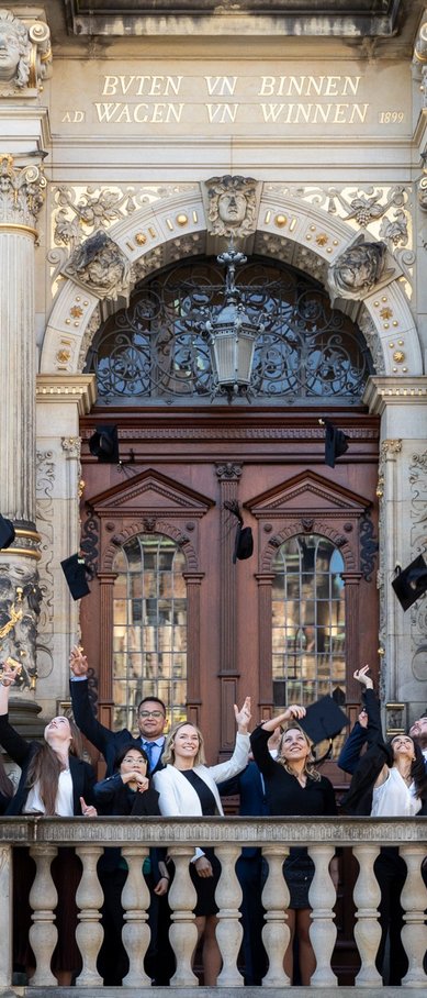 Gruppe von Absolventen in formeller Kleidung, die ihre Hüte vor einem historischen Gebäude in die Luft werfen.