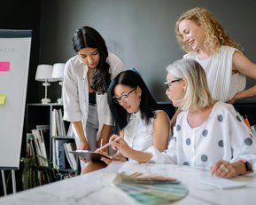 Four women are working together at a table while looking at a tablet. In the background, sticky notes and color palettes are visible.