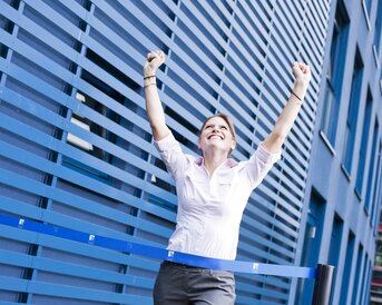 A woman in a white shirt raises her arms in the air and smiles as she crosses a finish line in front of a blue wall.