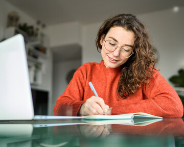 A young woman with curly hair and glasses is writing in a notebook while sitting at a table with a laptop.