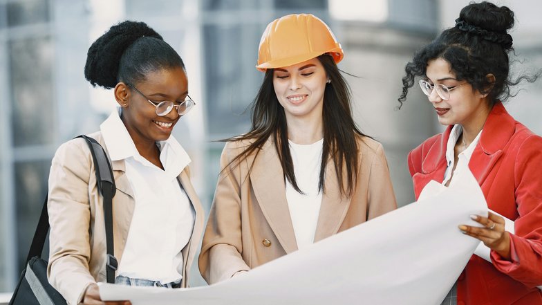 Drei Frauen betrachten gemeinsam einen Bauplan, eine trägt einen orangefarbenen Helm, während die anderen Notizen machen und diskutieren.