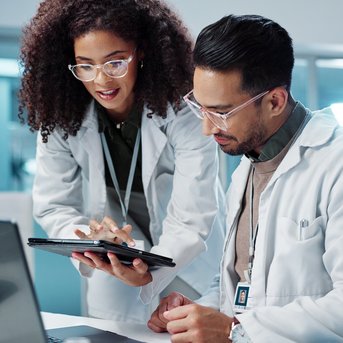 Zwei Wissenschaftler in Laborkitteln arbeiten gemeinsam an einem Laptop und einem Tablet in einem modernen Labor.