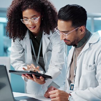 Two scientists in lab coats are working together on a laptop and a tablet in a modern laboratory.
