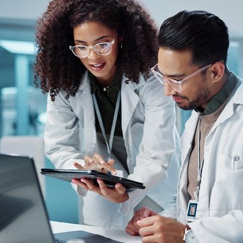 Two scientists in lab coats are working together on a laptop and a tablet in a modern laboratory.