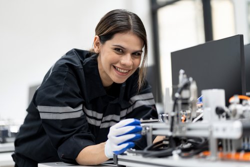 Eine junge Frau in Arbeitskleidung arbeitet an einer Maschine und hält ein Werkzeug in der Hand, während sie lächelt.