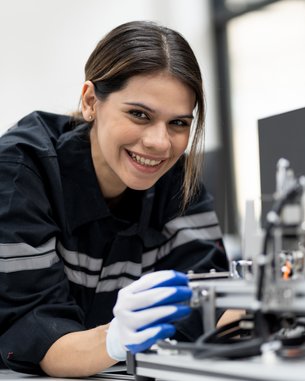 Eine junge Frau in Arbeitskleidung arbeitet an einer Maschine und hält ein Werkzeug in der Hand, während sie lächelt.
