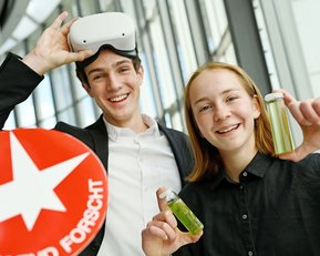 Zwei junge Menschen lächeln, einer trägt eine VR-Brille und hält ein Schild mit dem Text "Hier forscht". Die andere Person zeigt zwei Fläschchen.