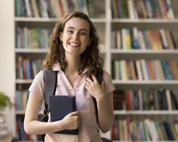 Eine junge Frau mit lockigem Haar trägt einen Rucksack und hält ein Buch in der Hand, lächelnd vor einem Bücherregal.