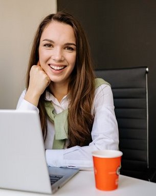 Eine lächelnde Frau mit langen Haaren sitzt an einem Schreibtisch, vor einem Laptop und einer Kaffeetasse, in einem modernen Büro.