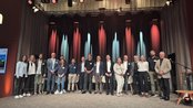 Eine Gruppe von Menschen steht auf einer Bühne, umgeben von bunten Lichtern. Sie lächeln und posieren für ein Foto während einer Veranstaltung.
