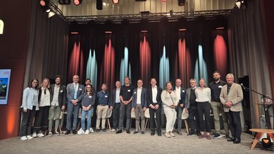 Eine Gruppe von Menschen steht auf einer Bühne, umgeben von bunten Lichtern. Sie lächeln und posieren für ein Foto während einer Veranstaltung.