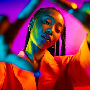 Eine Person mit Zöpfen trägt eine orangefarbene Jacke und posiert vor einem bunten, leuchtenden Hintergrund.