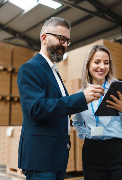 Ein Mann und eine Frau stehen in einem Lager, betrachten ein Tablet und lächeln. Im Hintergrund sind gestapelte Kartons sichtbar.