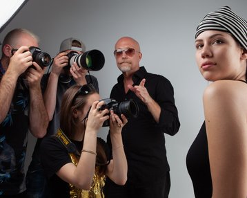 Eine Gruppe von Fotografen umringt ein Model, das in einem schwarzen Outfit und einem gestreiften Kopftuch posiert. Blitzlichter und Kameras sind sichtbar, während das Model in die Kamera schaut.