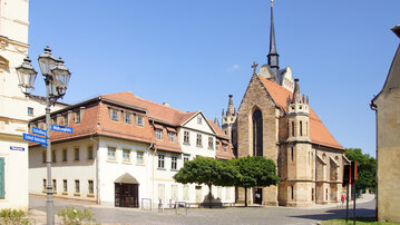Eine historische Straße mit einer Kirche im Hintergrund, umgeben von traditionellen Gebäuden und Bäumen. Der Himmel ist klar und blau, was eine ruhige Atmosphäre schafft.
