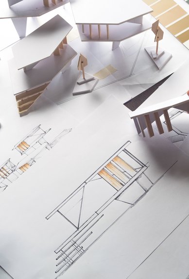 Eine Hand hält ein Modellhaus über Architekturzeichnungen und Farbmustern, die auf einem Tisch verteilt sind.