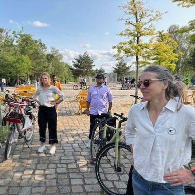 Eine Gruppe von vier Personen steht in einem Park, umgeben von Fahrrädern. Zwei Personen tragen lässige Kleidung, während eine Frau mit langen Haaren eine Sonnenbrille trägt. Im Hintergrund sind weitere Parkbesucher und Sitzgelegenheiten sichtbar.