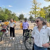 Eine Gruppe von vier Personen steht in einem Park, umgeben von Fahrrädern. Zwei Personen tragen lässige Kleidung, während eine Frau mit langen Haaren eine Sonnenbrille trägt. Im Hintergrund sind weitere Parkbesucher und Sitzgelegenheiten sichtbar.