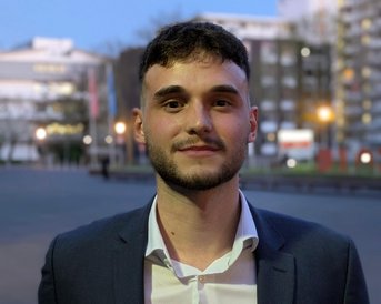 Ein junger Mann mit dunklem Haar trägt einen Anzug und hält eine Mappe in der Hand, während er vor einer urbanen Kulisse steht.