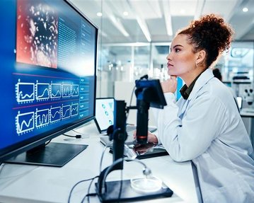 Eine Wissenschaftlerin in einem Labor betrachtet konzentriert Daten auf einem großen Bildschirm, während sie an einem Tisch sitzt.