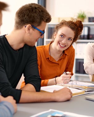 Vier junge Erwachsene sitzen an einem Tisch und diskutieren. Eine Frau lächelt, während die anderen aufmerksam zuhören. Bücher und Notizen liegen auf dem Tisch.