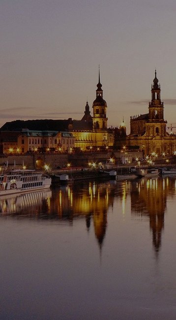 Die Silhouette der Stadt Dresden am Abend, mit historischen Gebäuden und Türmen entlang des Flusses Elbe. Mehrere Schiffe liegen am Ufer, während sich die Lichter der Stadt im Wasser spiegeln.