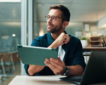 Ein Mann mit Brille sitzt an einem Tisch, hält ein Tablet und schaut nachdenklich. Im Hintergrund sind Bücherregale und Pflanzen sichtbar.