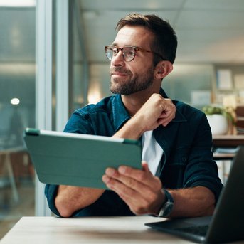 Ein Mann mit Brille sitzt an einem Tisch, hält ein Tablet und schaut nachdenklich. Im Hintergrund sind Bücherregale und Pflanzen sichtbar.