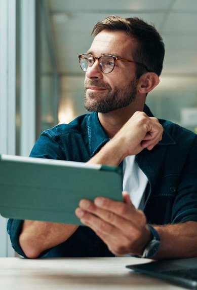 Ein Mann mit Brille sitzt an einem Tisch, hält ein Tablet und schaut nachdenklich. Im Hintergrund sind Bücherregale und Pflanzen sichtbar.