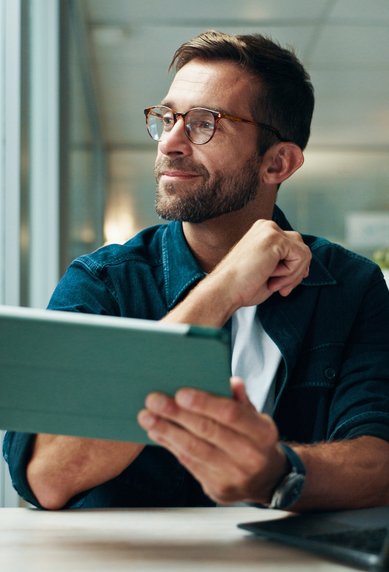 A man with glasses sits at a table, holding a tablet and looking thoughtfully. In the background, bookshelves and plants are visible.