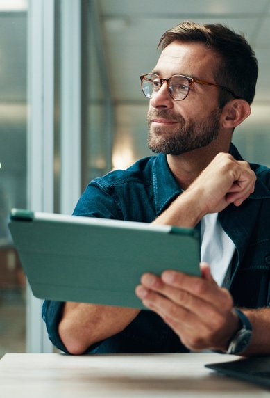 A man with glasses sits at a table, holding a tablet and looking thoughtfully. In the background, bookshelves and plants are visible.