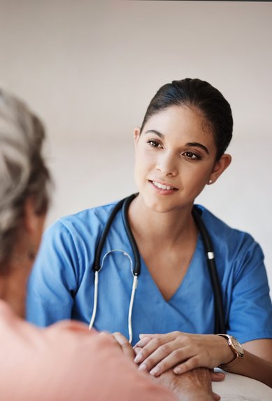 Eine Krankenschwester in blauer Uniform spricht mit einem älteren Patienten, der auf einem Stuhl sitzt.